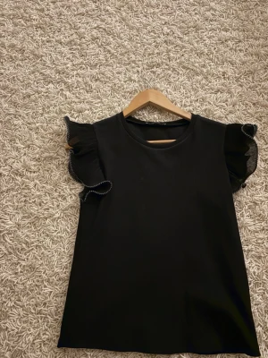 Svart topp med volangärmar - Snygg svart topp med plisserade volangärmar och dekorativ vit söm längs kanten. Toppen har rund halsringning och är tillverkad i ett mjukt tyg som ger en stilren look. Perfekt för dig som gillar trendiga detaljer och vill sticka ut lite extra.