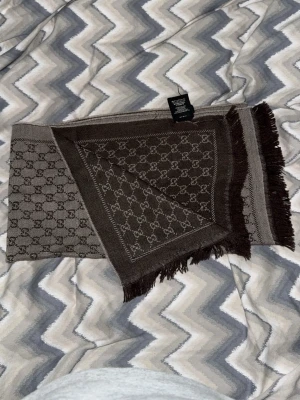 Gucci Halsduk - Snygg brun halsduk från Gucci med klassiskt GG-monogram i hela tyget. Halsduken har fransade kanter och är vävd i ett mjukt material som känns lyxigt. Perfekt accessoar för att lyfta din outfit.