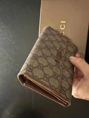 Gucci brun monogram plånbok - Snygg brun plånbok från Gucci med klassiskt GG-monogram i canvas och detaljer i brunt läder. Plånboken har en vikbar design och stilren metallogo framtill. Perfekt för dig som gillar lyxiga accessoarer med ikonisk design.   Vinted så har sina skavanker 