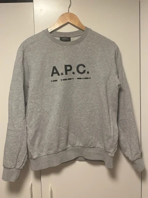 Grå sweatshirt från A.P.C. - Snygg grå sweatshirt från A.P.C. med svart logotyptryck på bröstet. Tröjan har rund halsringning, ribbade muddar vid ärmslut och nederkant samt en avslappnad passform. Perfekt för en clean och stilren look.