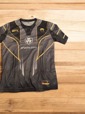 Svart och guld UFC t-shirt Venum - Snygg svart t-shirt från Venum i samarbete med UFC, med guld- och gråa detaljer samt tryck på bröstet och ärmarna. Materialet är lätt och andas, perfekt för träning eller sport. Stort UFC-tryck och logga framtill samt på ärmen. KAN KÄNNAS MINDRE FÖR DEN ÄR COMPRESSED