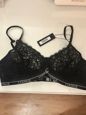 Svart spets-bh från Lounge - Snygg svart bh från Lounge med spetsdetaljer och elastiskt band med logga. Bh:n har tunna axelband och är tillverkad i ett genombrutet spetsmaterial som ger en feminin look. Perfekt för dig som gillar stilrena och bekväma underkläder. Helt oanvänd. Säljer pga att den är för liten. Nypris 525kr.