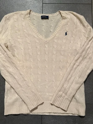 Kabelstickad vit tröja Polo Ralph Lauren - Kabelstickad vit tröja från Polo Ralph Lauren med V-ringning och lång ärm i 90% merinoull och 10% Kashmir . Klassisk design med broderad marinblå logga på bröstet och ribbade muddar. Perfekt för en stilren och tidlös look.