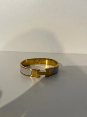 Hermes armband  - Snyggt armband i guldton med vit emalj och ett H-format spänne framtill. Armbandet har en stilren och modern design som passar perfekt till din vardagsstil. Metall och emalj ger en exklusiv känsla och den vita färgen gör det lätt att matcha med olika outfits.