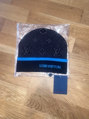 Svart mössa  - Snygg svart mössa med klassiskt monogrammönster och blå rand. Märkets logga är broderad framtill i blått. Mössan är stickad och har en modern, stilren look som passar perfekt till streetwear och modeintresserade.