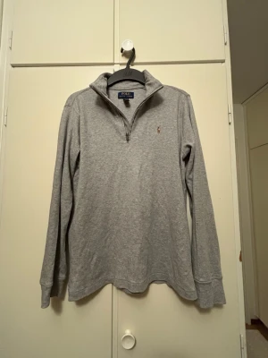 Grå halvzip tröja från Polo Ralph Lauren - Klassisk grå långärmad tröja från Polo Ralph Lauren med halv dragkedja och liten broderad logga på bröstet. Tillverkad i mjukt bomullsmaterial och har en avslappnad passform med ribbade muddar vid ärmslut och nederkant.