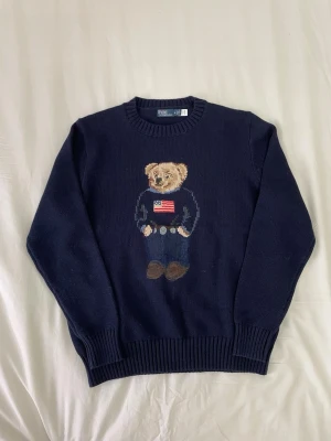 Mörkblå stickad tröja Polo Bear - Mörkblå stickad tröja från Polo Ralph Lauren med ikoniskt Polo Bear-motiv framtill. Rund halsringning och ribbade muddar. Tröjan har en klassisk passform och är tillverkad i mjukt material som känns skönt mot huden.