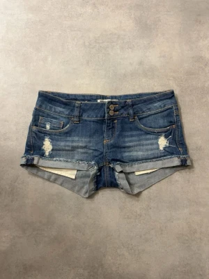 Blå jeansshorts med slitningar - Snygga blå jeansshorts med råa kanter och slitna detaljer framtill. Klassisk femficksmodell med dubbla knappar i midjan och uppvikta benslut. Perfekta för varma dagar och en avslappnad stil.