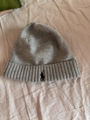 Grå mössa från Polo Ralph Lauren - Säljer en stilren grå mössa från Polo Ralph Lauren med uppvikt kant och den klassiska svarta logotypen broderad framtill. Mössan är ribbstickad i nederkant och tillverkad i mjukt stickat material som håller dig varm under kalla dagar. I nytt skick utan tagen har bara testat den en gång 