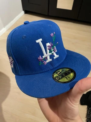 Blå LA Dodgers 59FIFTY keps med broderi - Snygg blå LA Dodgers 59FIFTY keps från New Era med vit LA-logga och lila blommor broderade på framsidan. På sidan finns ett lila och vitt 1988 World Series-märke. Undersidan av skärmen är ljuslila. Perfekt för dig som gillar streetwear och baseball. storlek 7 1/8 eller 56,8 cm. 