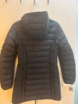 Svart dunjacka med huva Parajumpers - Jackan är en svart, lång dunjacka från Parajumpers gjord i ett superlätt material. Den har huva, dragkedja framtill och en diskret logga på ärmen. Insidan är ljusgrå och har även en inne ficka och jackan är utmärkt för kalla vinterdagar/vår dagar. Stilen är både snygg och stilren. Jackan är i mycket bra skick och köptes för 5599 kr. Den har även 4 stora ute fickor med dragkedja Varumärke Parajumpers Storlek XS Skick Mycket bra Material Dun