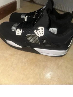Svarta Air Jordan 4 Retro sneakers - Säljer ett par svarta Air Jordan 4 Retro sneakers med vita och grå detaljer. Skorna har meshpaneler på sidorna, klassisk Jumpman-logga på plösen och häl, samt svarta skosnören. Ovandelen är i syntetmaterial och mockaliknande textur.