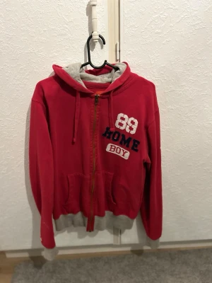 Röd hoodie från Homeboy med tryck - Röd hoodie från Homeboy med dragkedja, grå huva och broderat tryck med siffran 89 och texten HOME BOY på bröstet. Mjuk insida och ribbade muddar i grått. Perfekt för en avslappnad streetwear-look.