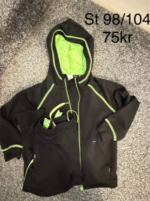 Svart träningsset med limegröna detaljer - Säljer ett svart träningsset med hoodie och byxor, båda med limegröna sömmar och dragkedja. Hoodien har meshfoder i huvan och praktiska fickor. Perfekt för sport eller fritid.