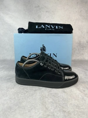 Svarta Lanvin sneakers i mocka och lack - Stilrena svarta sneakers från Lanvin med ovandel i mjuk mocka och glansig lackad tå. Skorna har svarta skosnören, metallfärgade öljetter och en svart sula. Perfekta för dig som gillar en clean och lyxig look.
