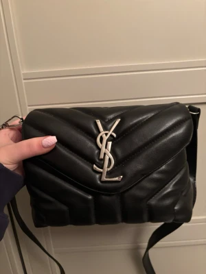 Svart axelväska från Saint Laurent - Säljer en svart quiltad axelväska från Saint Laurent med ikoniskt YSL-emblem i silver på framsidan. Väskan är tillverkad i mjukt läder och har justerbar axelrem. Perfekt för dig som vill ha en stilren och lyxig accessoar.