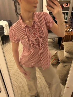Rödvit randig skjorta med volang - Säljer en rödvit randig skjorta med knyt framtill, resår på baksidan och knappar hela vägen. Skjortan har korta ärmar, klassisk krage och är gjord i ett lätt bomullstyg. Perfekt för dig som gillar en söt och trendig look. Superfin❤️