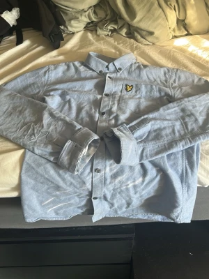 Ljusblå skjorta från Lyle & Scott - Snygg ljusblå skjorta från Lyle & Scott Junior med klassisk krage och knappstängning framtill. Skjortan har lång ärm och den ikoniska gula loggan på bröstet. Perfekt till både jeans och chinos för en clean look.