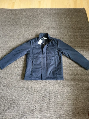 Overshirt - Passar M. Snygg mörkblå vindjacka från Arket med ståkrage och dragkedja samt tryckknappar framtill. Jackan har två bröstfickor med lock och två sidofickor. Tillverkad i ett slätt, vindtätt material som passar perfekt till blåsiga dagar.