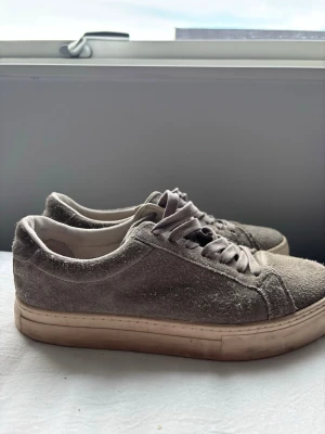 Grå mockasneakers med vit sula - Snygga sneakers i grå mocka med klassisk låg profil och vita sulor. Skorna har matchande grå skosnören och en stilren design som passar till det mesta. Perfekta för dig som gillar en clean och enkel look.