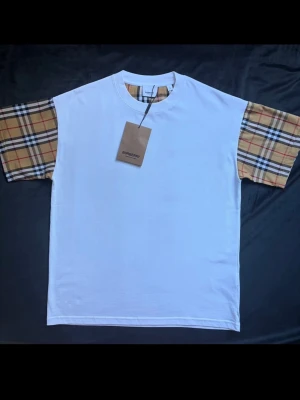  Burberry t-shirt  - Snygg vit t-shirt från Burberry med klassiska rutiga ärmar. Avslappnad passform storlek S men passar mera som M. Perfekt för dig som vill sticka ut nu på sommaren