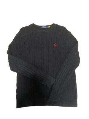 Svart kabelstickad tröja Polo Ralph Lauren - Kabelstickad svart tröja från Polo Ralph Lauren med rund halsringning och röd broderad logga på bröstet. Tröjan har ribbade muddar vid ärmslut och nederkant. Perfekt för en stilren och klassisk look.
