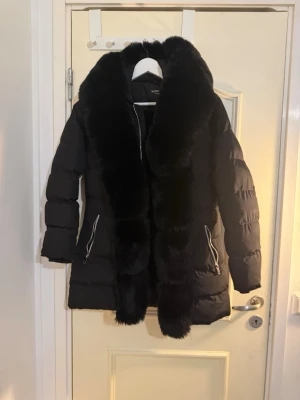 Svart dunjacka med fuskpäls - Stilren svart dunjacka från Marzi med stor, mjuk fuskpäls längs krage och dragkedja. Jackan har två snedställda fickor med dragkedja och quiltad design. Perfekt för kalla vinterdagar och ger en lyxig känsla till din outfit.