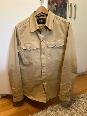 Beige overshirt från Oscar Jacobson - Stilren beige overshirt från Oscar Jacobson med klassisk krage, två bröstfickor med lock och knappar framtill. Overshirten har raka ärmar med manschett och är tillverkad i ett kraftigare bomullstyg. Perfekt för lager-på-lager-stil.