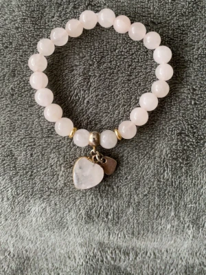 Armband i rosenkvarts - Ett rosenkvarts-armband med lite guldpärlor och hjärtberlocker på. Armbandet är köpt på Via Brazil och är i väldigt bra skick. Stenen rosenkvarts är känd för att vara kärlekens sten, men den fokuserar mest på att man ska älska sig själv. Rosenkvarts står även för självförtroende, balans, lugn och harmoni.