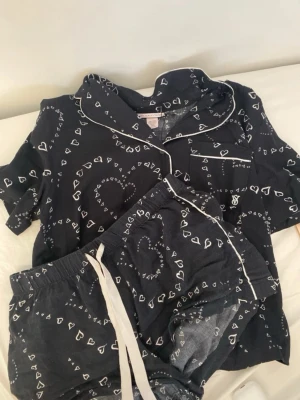 Svart pyjamas-set Victoria's Secret - Sött svart pyjamas-set från Victoria's Secret med vita hjärtmönster och vita kanter. Setet består av en kortärmad skjorta med knappstängning och bröstficka samt matchande shorts med dragsko i midjan. Perfekt för mysiga kvällar hemma.