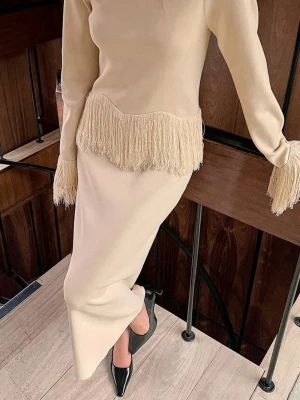 Beige långklänning med fransar - Stilren beige långklänning med fransdetaljer vid midjan och ärmsluten. Klänningen har lång ärm och rak passform som ger en elegant siluett. Perfekt för dig som vill sticka ut med en unik detalj. Matcha med klackar för en extra snygg look.