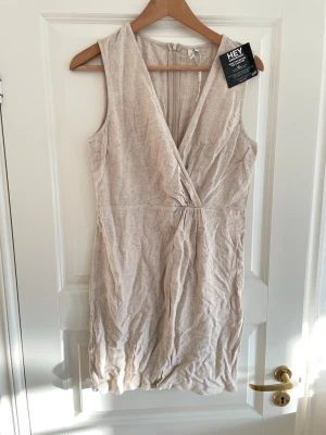 Beige omlottklänning i klänning  - Snygg beige omlottklänning från HEY i linne. Ärmlös modell med v-ringning och dragkedja i ryggen. Perfekt för varma dagar och enkel att styla med accessoarer.