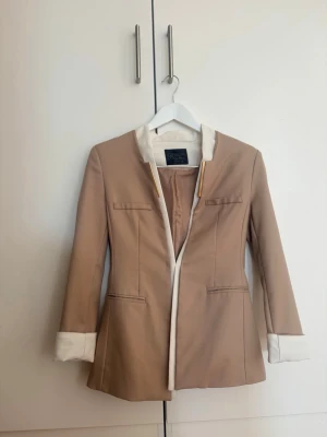 Beige kavaj med vita detaljer Bling Bling - Stilren beige kavaj från Bling Bling med vita kontrastdetaljer på krage och ärmslut. Långärmad modell med markerade axlar och dekorativa fickor framtill. Guldiga detaljer vid kragen ger en extra touch. Perfekt för en trendig och elegant look.