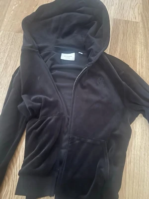 Svart velourhoodie från Juicy Couture - Mjuk och lyxig svart hoodie i velour från Juicy Couture med dragkedja framtill och broderad JC-logga på bröstet. Klassisk huva och två fickor framtill. Perfekt för en chill och trendig look. Bra skick 