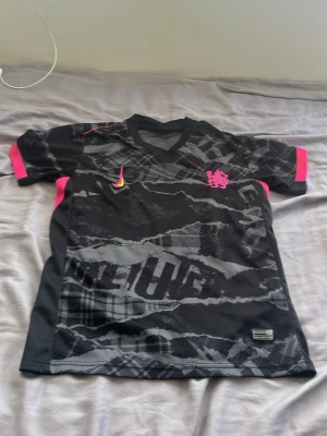 Nike svart och rosa fotbollströja - Snygg svart fotbollströja från Chelsea och Nike med grått och svart mönster, rosa detaljer på ärmar och axlar samt rosa klubbmärke på bröstet. Tröjan har en gul Nike-logga och är tillverkad i ett lätt, andningsbart material perfekt för träning.