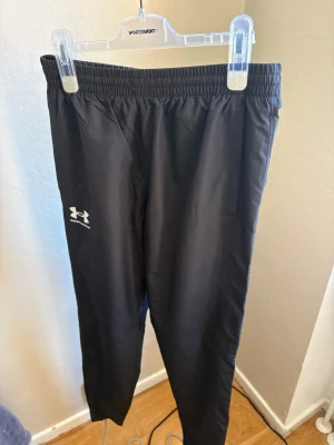 Svarta träningsbyxor Under Armour - Säljer ett par svarta träningsbyxor från Under Armour med elastisk midja och logga på vänster ben. Tillverkade i ett lätt och snabbtorkande material som passar perfekt för löpning eller gym. Snygg och enkel design med smal passform.