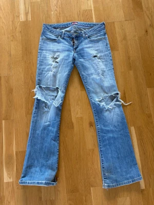 Ljusblå slitna bootcut jeans - Säljer ett par lågmidjade ljusblå bootcut jeans med stora slitningar och hål över knäna. (Gylfen har gått sönder men du som är bra på att sy, kan fixa det lätt!!)