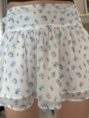 Vit blommig kortkjol med volang - Fluffig vit kortkjol med blått blommönster och volangkant. Kjolen har bred linning och är gjord i ett tunt, luftigt material som ger en flowy känsla. Perfekt för sommar och vår när du vill ha en söt och romantisk look.