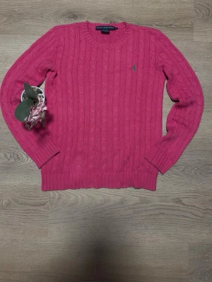 Rosa kabelstickad tröja Ralph Lauren - Snygg rosa kabelstickad tröja från Ralph Lauren med rund halsringning och diskret broderad logga på bröstet. Tröjan har långa ärmar och ribbade muddar vid ärmslut och nederkant. Perfekt för dig som gillar klassisk och stilren design. DET STÅR S MEN DE PASSAF M
