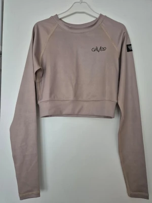 Träningströja - Säljer en ljusbeige croppad träningströja långärmad från Gavelo med rund hals och diskret logga på bröstet. Toppen har en tight passform och är tillverkad i syntetmaterial som passar perfekt för träning. Snygga sömdetaljer och enkel design.