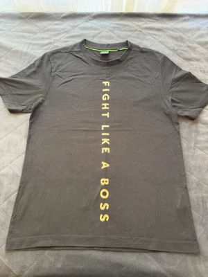 Svart BOSS t-shirt med tryck - Snygg svart t-shirt från BOSS med texten 'FIGHT LIKE A BOSS' i guld tryck längs framsidan. Klassisk rund hals och korta ärmar. Perfekt för dig som vill ha en enkel men statement t-shirt. Materialet känns mjukt och skönt mot huden.