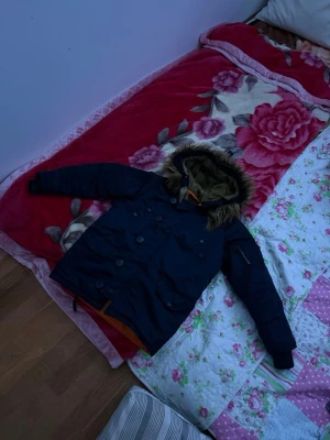 Mörkblå parkas med fuskpäls - Snygg mörkblå parkas med avtagbar huva och fuskpälsdetalj. Jackan har dubbelknäppning framtill, flera fickor och orange innerfoder. Perfekt för kalla dagar och ger en cool streetstil.