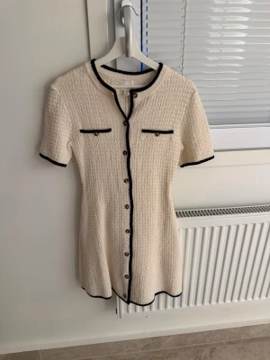 Vit kortklänning med svarta detaljer från hm - Stilren vit kortklänning med svarta kanter och dekorativa knappar framtill. Klänningen har korta ärmar, rund halsringning och två fejkfickor på bröstet. Materialet är strukturerat och ger en exklusiv känsla.