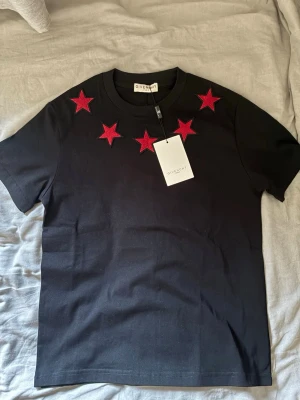 Svart t-shirt med röda stjärnor Givenchy - Svart t-shirt från Givenchy med röda stjärnor broderade runt halsringningen både fram och bak. Klassisk passform och korta ärmar. Tillverkad i mjuk bomull för en bekväm känsla. Perfekt för dig som vill ha en statement t-shirt med exklusiv känsla.