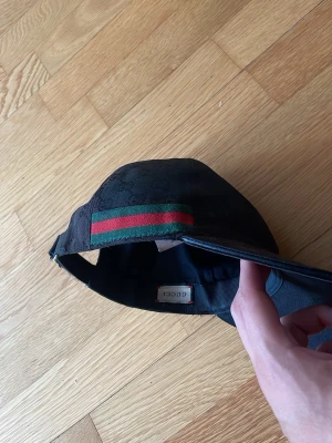 Svart Gucci keps med GG-mönster - Säljer en svart keps från Gucci med klassiskt GG-mönster över hela kepsen och den ikoniska gröna och röda randen på sidan. Justerbar rem bak och böjd skärm. Materialet är canvas med detaljer i läder och bomull. Perfekt för dig som vill ha en stilren och lyxig accessoar.
