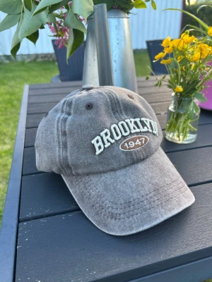 Brun keps med BROOKLYN 1947 brodyr - Säljer en brun keps med böjd skärm och broderad text 'BROOKLYN 1947' framtill. Kepsen har en avslappnad vintage-look och är tillverkad i bomullstyg. Perfekt för dig som gillar streetwear och vill ha en stilren accessoar.