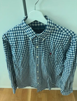 Blårutig skjorta från Ralph Lauren - Klassisk blårutig skjorta från Ralph Lauren med vit bas och blå rutor. Skjortan har knappar framtill, lång ärm och den ikoniska röda broderade loggan på bröstet. Tillverkad i mjuk bomull och perfekt för en clean och stilren look.