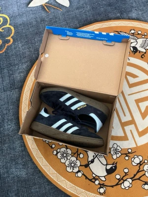 Adidas Handball Spezial marinblå - Snygga Adidas Handball Spezial sneakers i marinblå mocka med klassiska blå  ränder och brun gummisula. Guldfärgad Adidas-text på sidan och retrovibbar. Perfekta för dig som gillar streetwear och sportig stil. Storlek 38. Köpt för 1200 knappt använda.