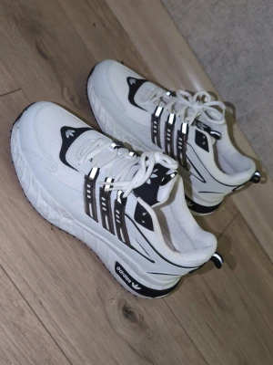 Vita Adidas sneakers med svarta detaljer - Snygga vita sneakers från Adidas med svarta detaljer och klassiska tre ränder på sidan. Skorna har en chunky sula, mesh- och syntetmaterial samt logga på plösen och hälen. Perfekta för dig som gillar streetwear och sportig stil. Helt nya st 42 riktigt  snygga skor for real :nypris  2900