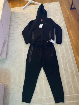 Svart Nike set med dragkedja - Snygg svart hoodie från Nike med dragkedja framtill och två fickor med dragkedja. Klassisk Nike-logga på bröstet och huva med snörning. Tillverkad i mjukt material som passar perfekt till en avslappnad stil.